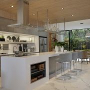 A luxury open-plan kitchen design contrasting warm wood ceilings and marble floors with clean, minimalist Silestone Iconic White worktops